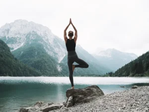 Person practicing yoga in tree pose on a rocky lakeshore with mountains in the background