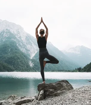 Person practicing yoga in tree pose on a rocky lakeshore with mountains in the background