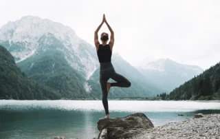 Person practicing yoga in tree pose on a rocky lakeshore with mountains in the background
