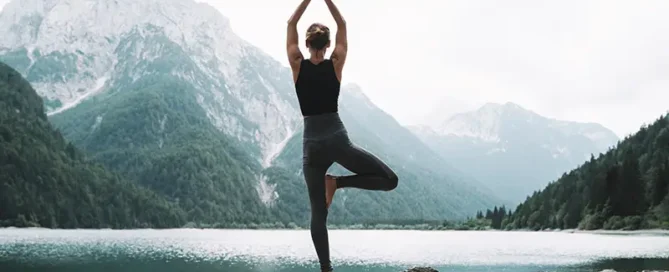 Person practicing yoga in tree pose on a rocky lakeshore with mountains in the background