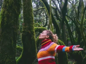 Woman in a bright striped sweater stands with arms outstretched in a mossy forest, eyes closed and enjoying nature.