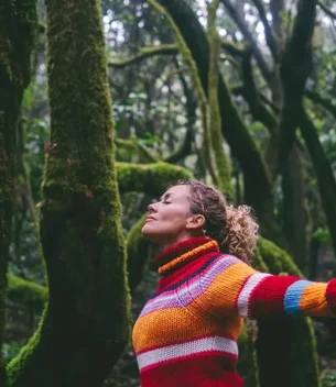 Woman in a bright striped sweater stands with arms outstretched in a mossy forest, eyes closed and enjoying nature.