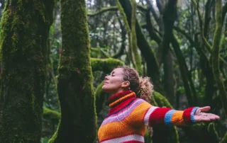 Woman in a bright striped sweater stands with arms outstretched in a mossy forest, eyes closed and enjoying nature.