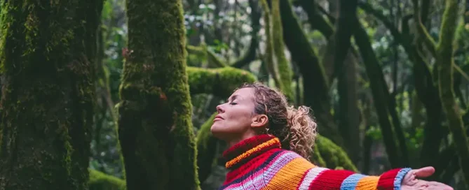 Woman in a bright striped sweater stands with arms outstretched in a mossy forest, eyes closed and enjoying nature.