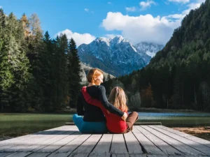 Two people sit on a wooden dock by a lake, arms around each other, with forested mountains in the background.