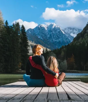 Two people sit on a wooden dock by a lake, arms around each other, with forested mountains in the background.