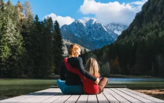 Two people sit on a wooden dock by a lake, arms around each other, with forested mountains in the background.