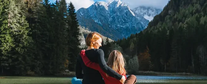 Two people sit on a wooden dock by a lake, arms around each other, with forested mountains in the background.