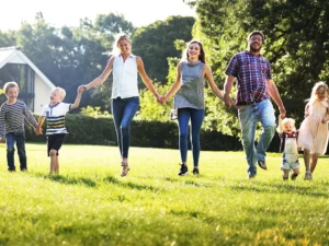 Group of seven people holding hands and running across a sunny park lawn (three adults and four children).