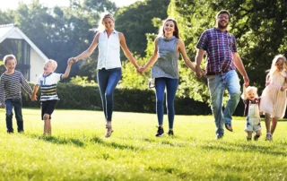 Group of seven people holding hands and running across a sunny park lawn (three adults and four children).