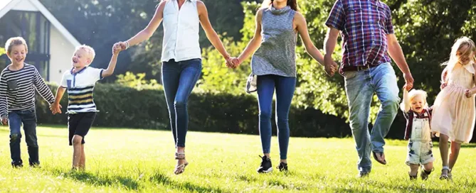 Group of seven people holding hands and running across a sunny park lawn (three adults and four children).