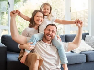 Dad lifts his daughter on his shoulders while mom smiles beside them in a bright living room. Contains arms up and happy faces on a couch setup.