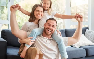 Dad lifts his daughter on his shoulders while mom smiles beside them in a bright living room. Contains arms up and happy faces on a couch setup.