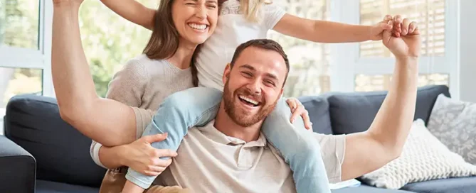 Dad lifts his daughter on his shoulders while mom smiles beside them in a bright living room. Contains arms up and happy faces on a couch setup.