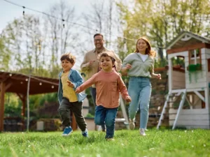 A family of four runs across a sunny lawn, with two children leading and adults chasing behind, smiling as they play outside near a wooden playhouse.