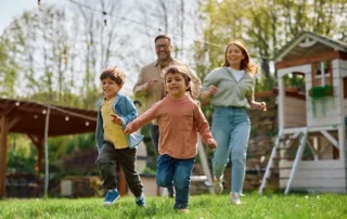 A family of four runs across a sunny lawn, with two children leading and adults chasing behind, smiling as they play outside near a wooden playhouse.