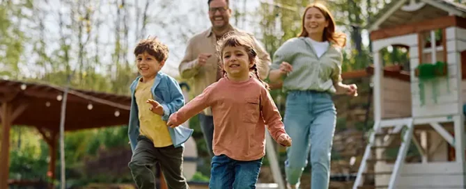 A family of four runs across a sunny lawn, with two children leading and adults chasing behind, smiling as they play outside near a wooden playhouse.